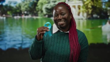 Parkta elinde mavi kurdeleyle gülümseyen bir kadın. Canlı yeşillik suya yansıyor. Açık havada barışçıl farkındalığı simgeliyor..