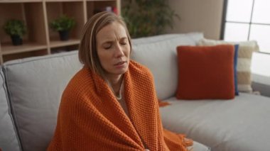 Turuncu battaniyeye sarılmış bir kadın rahat bir oturma odasında, yumuşak yastıklar ve ahşap raflarla sıcak bir ev atmosferinde keyifsiz ve yorgun görünüyor..