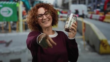 İnşaat alanında elinde dolarlarla gülümseyen bir kadın, açık havada dururken işaret ederek, başarılı bir bağış toplama veya tasarruf çabası sergiliyor..