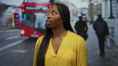 Şehir merkezinde otobüs durağında dikilen, arka planda kırmızı çift katlı otobüsü olan sarı tişörtlü bir kadın.