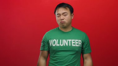 Gönüllü metin içeren yeşil tişört giyen genç bir adam kızgın bir ifadeyi gösteren kırmızı bir arka plana karşı kollarını kavuşturmuş duruyor..