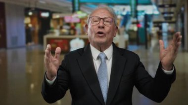 İş elbisesi giymiş kıdemli bir adam alışveriş merkezinin içinde el kol hareketi yapıyor. Görünür bir merkez mağazası. Bulanık alışveriş geçmişi olan profesyonel bir ev ortamını canlandırıyor..