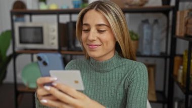 Ofis ortamında akıllı telefon ve kredi kartı kullanan yeşil kazaklı bir kadın modern teknoloji ve finans ürünleri sergiliyor..