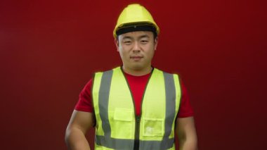 İnşaat işçisi koyu kırmızı arka planda anlamlı bir jest yapıyor sarı kaskını ve yansıtıcı yeleğini vurguluyor..