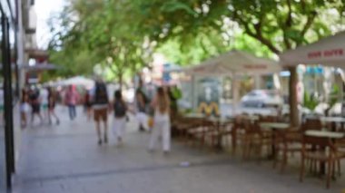 Bulanık insanlar, güneşli bir günde bokeh kafeler ve ağaçlarla açık bir sokakta yürüyorlar. Dağınık ve canlı bir şehir atmosferi yaratıyorlar.