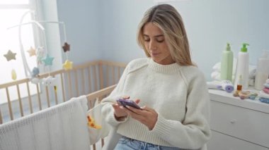 Bebek beşiği ve oyuncaklarıyla çevrili akıllı telefonu olan bir kadın yatak odasında oturuyor. Bakıcı ve huzurlu bir ortam yaratıyor..