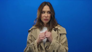 Mavi arka plan üzerinde trençkotlu kadın. Düşünceli bir yüz ifadesi var. Tecrit edilmiş ve odaklanmış.