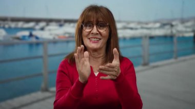 Sahildeki limanda arka planda teknelerle el çırpan bir kadın açık hava ortamında neşe saçıyor.