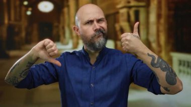 Mavi gömlekli kel bir adam kapalı bir kilisede parmak uçlarında çelişkili duygular ve dövmeler sergiliyor bulanık bir arkaplanda..