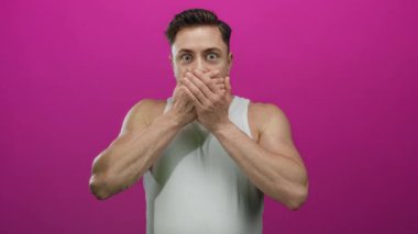 Sakallı genç adam elleriyle ağzını kapatıyor, izole bir ortamda canlı pembe duvara karşı duruyor, vücut dili ve yüz ifadesi ile şaşkınlık ve şaşkınlık ifade ediyor..