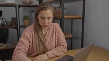 Pembe kazaklı kadın ofiste dizüstü bilgisayarda otururken stresli görünüyor. Arka planda raflar işyeri ayarlarını gösteriyor..