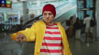 Havaalanı terminalinde sarı bir yağmurluk ve kırmızı bir bere takmış, kapalı bir alanda hareketli hareketler yapan genç İspanyol bir adam..