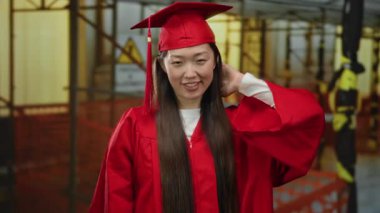 Kırmızı mezuniyet elbisesi giymiş bir kadın inşaat alanında kendine güvenerek gülümsüyor. Güzelliği ve akademik başarıyı somutlaştırıyor. Endüstriyel bir arka planda..