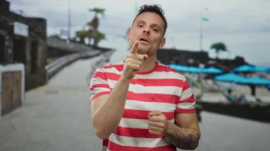 Kumsalda güneşli bir havada mavi şemsiyeler altında çizgili gömlek ve dövmeli yakışıklı bir adam.