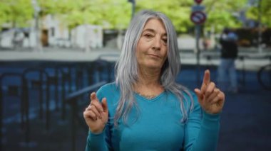Gri saçlı yaşlı bir kadın işlek bir caddede bir jest yapıyor, canlı şehir ortamını yakalıyor..