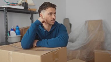 Yeni dairenin mukavva kutusuna yaslanmış mavi kapüşonlu genç adam paketlenmemiş eşyalarla çevrili modern yerleşim alanındaki geçişi ve yeni başlangıçları sembolize ediyor..