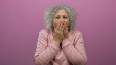 Kıvırcık gri saçlı yaşlı bir kadın ağzını canlı pembe bir duvara karşı şaşkınlıkla kapatıyor. Aynı süveteri giyiyor. Şok ve duygu ifadelerini somutlaştırıyor..