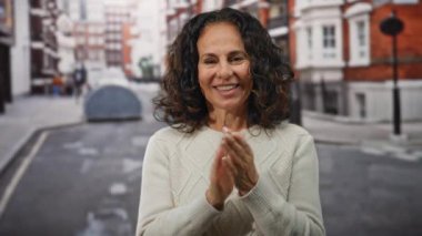 Kıvırcık saçlarıyla gülümseyen bir kadın şehir arka planında bulanık bir şekilde mutlu bir şekilde el çırpıyor, örgü örmüş bir kazak giyiyor, pozitif bir ifade ve neşeli bir tavır sergiliyor..