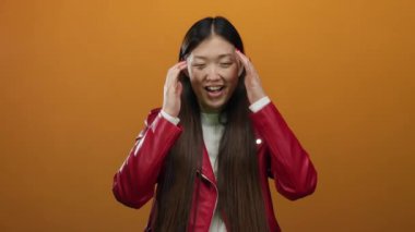 Yüzüne yakın gülümseyen bir kadın canlı turuncu bir arka plan karşısında, şık kırmızı bir ceket giyiyor ve neşe ve heyecan ifade ediyor..