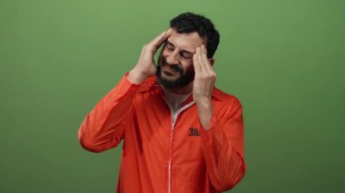 Turuncu tulumlu İspanyol sakallı adam üzgün ve dalgın görünen yeşil arka plana karşı duruyor..