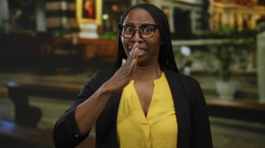 Şehir ortamında gözlüklü ve sarı gömlekli bir kadın, el hareketi ve Afro-Amerikan özellikleriyle şehir ortamında duygularını ifade ederken şaşırdı..