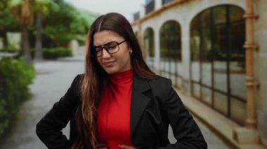 Yeşil ve kemerli mimariyle çevrili kentsel bir ortamda, gözlüklü ve kırmızı balıkçı yakalı bir yaka giyen bir kadın dışarıda acı dolu bir ifadeyle dikiliyor..