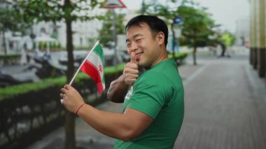 Başparmaklarıyla gülümseyen genç adam İran bayrağını şehir caddesinde tutuyor.