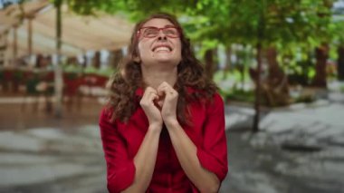 Kırmızı gözlüklü ve gömlekli bir kadın yeşil ağaçlar ve bulanık binalarla çevrili güneşli bir parkta dua ediyor..