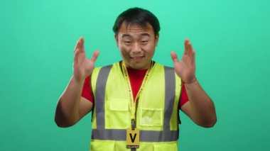 Sarı yelekli genç adam el işaretleriyle yeşil arka planı imzalıyor neşeli bir gülümsemeyle ifadesini gösteriyor.