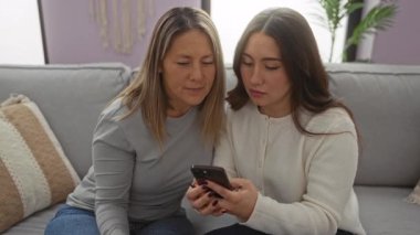 Anne ve kızı kanepede kaynaşıyor, rahat bir oturma odasında akıllı bir telefon kullanıyor, kapalı bir ev ortamında bir aile birlikteliği ve sevgi anı yakalıyor..