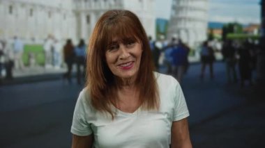 İspanyol kıdemli kadın pisa yakınlarında gülümsüyor, arka planda ikonik kuleyle güneşli bir günde şehir hayatının tadını çıkarıyor, gelişigüzel giyiniyor, kültürel çeşitlilik ve pozitiflik sergiliyor..