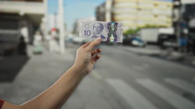 Arka planda şehir yaşamını gözler önüne seren bulanık binaları ve araçları olan işlek bir şehir caddesinde Kanada doları banknotu tutan adam..