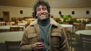 Kapalı bir restoranda kahve içerken gülümseyen genç bir adam, rahat ceketini ve neşeli ifadesini vurguluyor..