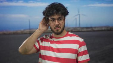 Sakallı, gözlüklü ve çizgili tişörtlü genç adam açık mavi gökyüzünün altındaki bir tarlada yel değirmenlerinin yanında el kol hareketleri yapıyor..