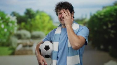 Parkta elinde futbol topu olan İspanyol bir adam, güneşli bir günde çevresi yeşilliklerle çevriliyken neşeli bir ifadeyle duygularını gösteriyor..