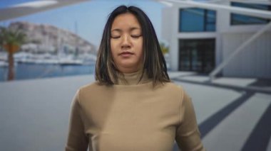 Bej renkli kazaklı bir kadın modern deniz kenarı binasının önünde ellerini sımsıkı tutarak limana yanaşmış teknelerle deniz kenarında yalvarışlarını gösteriyor..