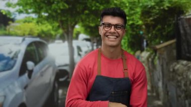 Dişlerine işaret eden gülümseyen gözlüklü ve önlüklü genç adam arka planda ağaçlar ve arabalarla dolu canlı bir şehir caddesinde duruyor..