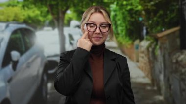 Canlı bir şehir caddesinde gözlük takan ve açık havada, yemyeşil ağaçlarla kaplı arka planda siyah bir ceket giyen bir kadın gözlerini işaret ediyor..