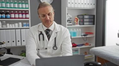 Klinikte gözlüklü, önlüklü, dizüstü bilgisayarlı ve rafları tıbbi malzemelerle dolu bir masada oturan olgun bir adam..