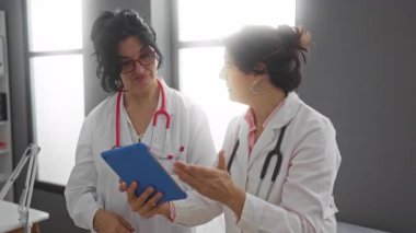 İki kadın doktor, bir hastane odasında, beyaz önlük ve stetoskop giyip, hasta verilerini tartışıyorlar.