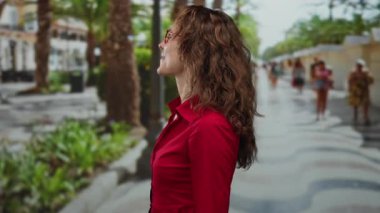 Kırmızı tişörtlü, gözlüklü, gülümseyen ve sunum yapan bir kadın park yolunda palmiye ağaçları ve arka plandaki insanlarla gündüz vakti dikilirken..