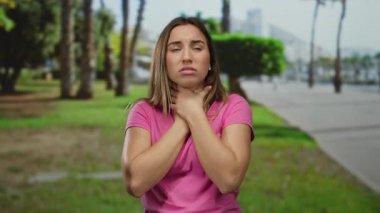 Pembe gömlekli genç bir kadın açık bir gökyüzünün altında ağaçlarla el ele tutuşarak yeşil manzara ve açık hava yollarıyla çevrili, rahatsızlık ve endişeyi gösteriyor..