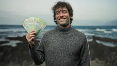 Deniz kenarında roubles tutan bir adam, arkasında okyanus dalgaları olan bulutlu bir gökyüzü altında mutluluk ve zenginlik sergiliyor.