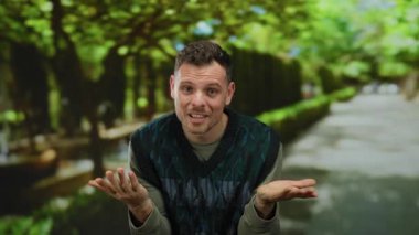 Adam dışarıda omuz silkiyor yeşil parkı günlük kıyafet ağaçlarını bulanıklaştırıyor arka planı bulanıklaştırıyor düşünceli ruh hali samimi portre duyguları doğal ortam sakinliği.