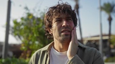 Kıvırcık saçlı ve sakallı genç adam parlak bir gökyüzünün altındaki şehir parkında yanağını tutarken düşünceli görünüyor.