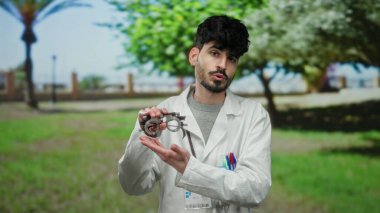 Gözlüklü ve laboratuvar önlüklü genç adam güneşli bir park ortamında optometri ekipmanı tutuyor. Profesyonel tıbbi davranışları doğal yeşillikle harmanlıyor..
