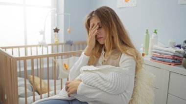 Sıcak yatak odasında yorgun görünen, beşiği yumuşak ışıkla çevrili, sessiz bir evde bebeği tutan bir kadın..