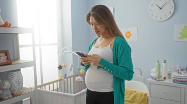 Bebek beşiğinin yanındaki yatak odasında akıllı telefon kullanan bir kadın, modern ve rahat bir ev tasarımıyla huzurlu, hamile bir atmosfer sergiliyor..