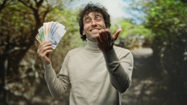 İspanyol adam, güneşli bir park ortamında Güney Kore banknotlarını tutarken neşeli bir şekilde kameraya öpücük yolluyor, canlı ve eğlenceli bir açık hava yaratıyor..