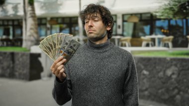 Şehir sokaklarında İspanyol bir adam Amerikan dolarlarını elinde tutuyor, Birleşik Devletler banknotlarını kendinden emin bir şekilde sergiliyor, finansal başarı hissi veriyor..